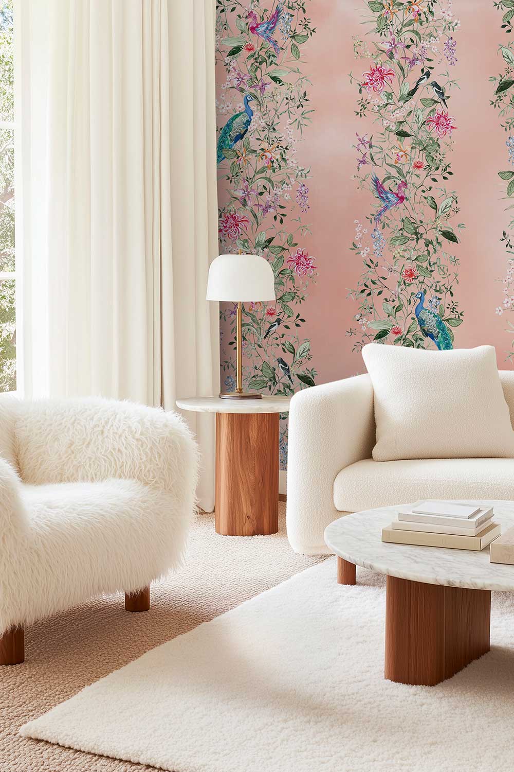 A styled living room corner featuring a blush pink wallpaper with a detailed botanical and bird pattern, paired with a white faux fur armchair, a cream sofa, and a marble-top coffee table. Hand-painted Mahjong Garden Stripe wallpaper featuring watercolor chinoiserie florals, peacocks, birds, bamboo, and botanical stripes on an blush pink coral background