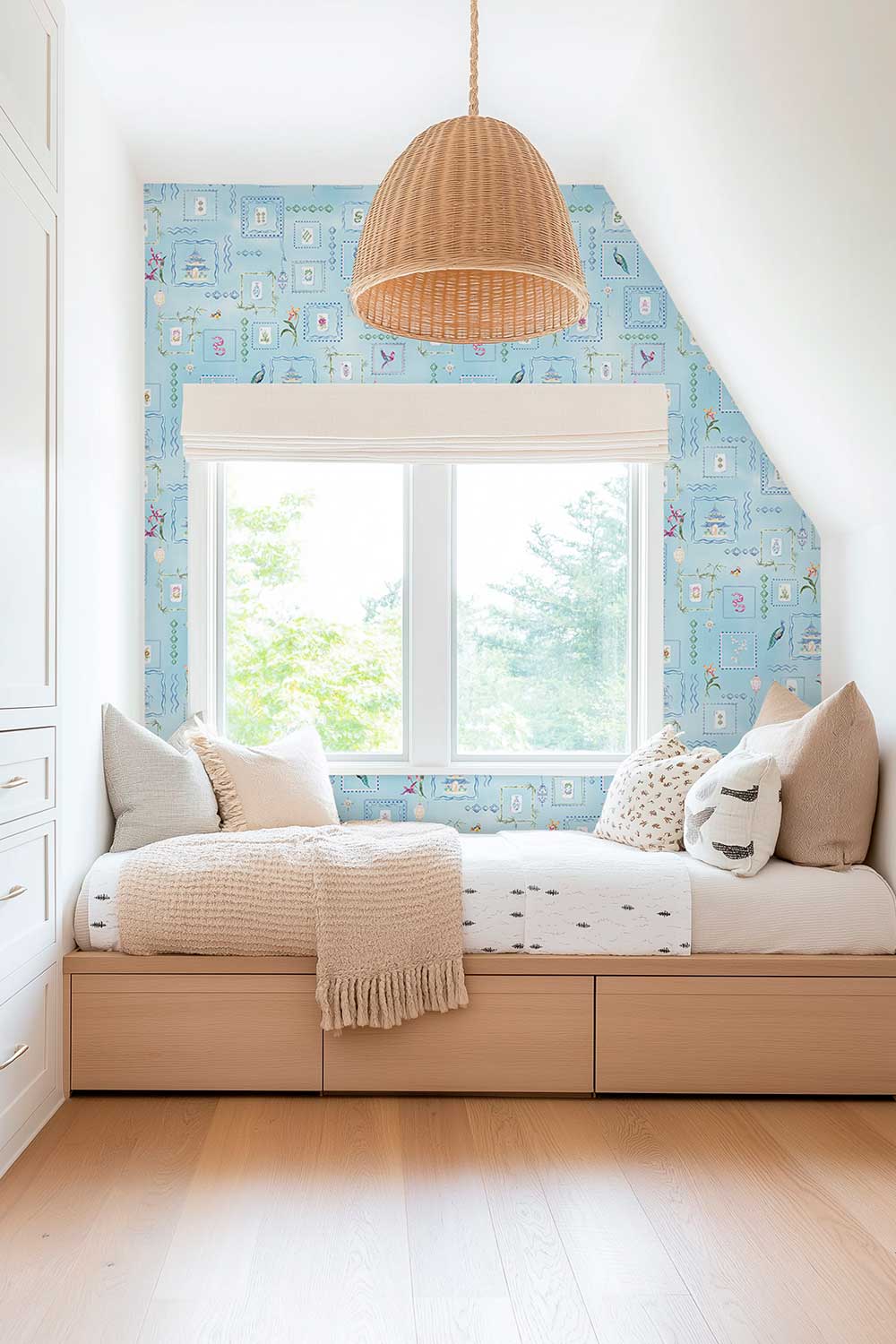 A styled children's room corner featuring a light wood platform daybed with white bedding and pillows, set against a blue patterned wallpaper and an angled ceiling. A natural wicker pendant light hangs above the bed. Soft blue Mahjong Tiles traditional matte wallpaper featuring hand-painted pastel watercolor mahjong tiles, pagodas, bamboo, ginger jars, birds, lanterns, and floral chinoiserie details.