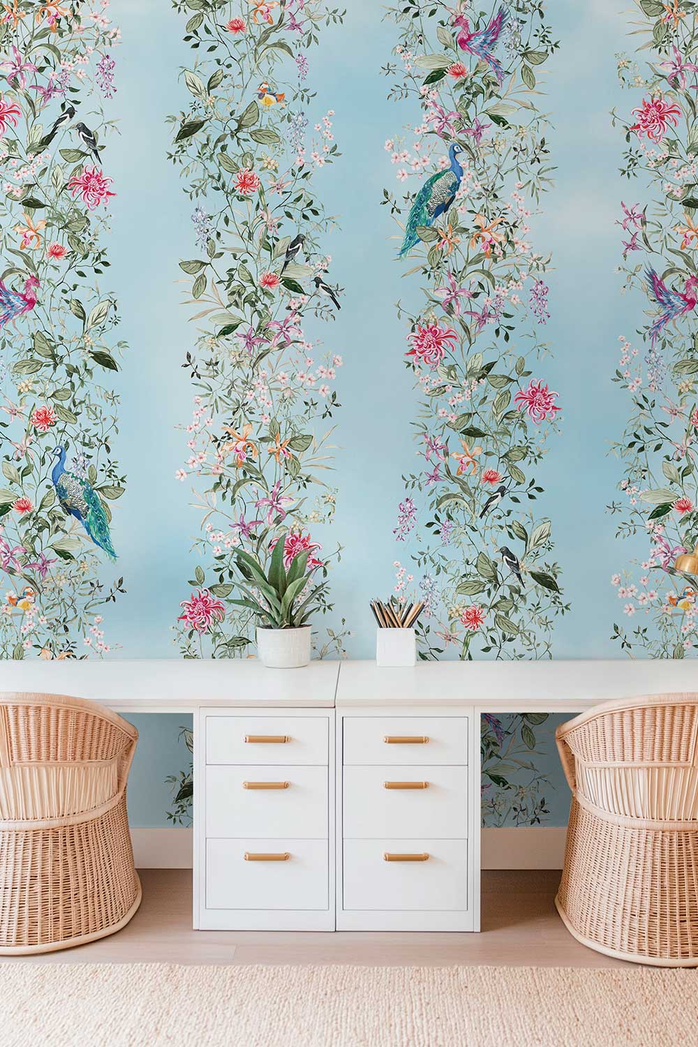 A styled interior scene featuring light blue wallpaper with vertical floral and bird motifs, a white bench with drawers, and two woven rattan chairs on a light rug. Natural grasscloth wallpaper featuring hand-painted Mahjong Garden Stripe with chinoiserie florals, peacocks, birds, bamboo, and botanical watercolor artwork.