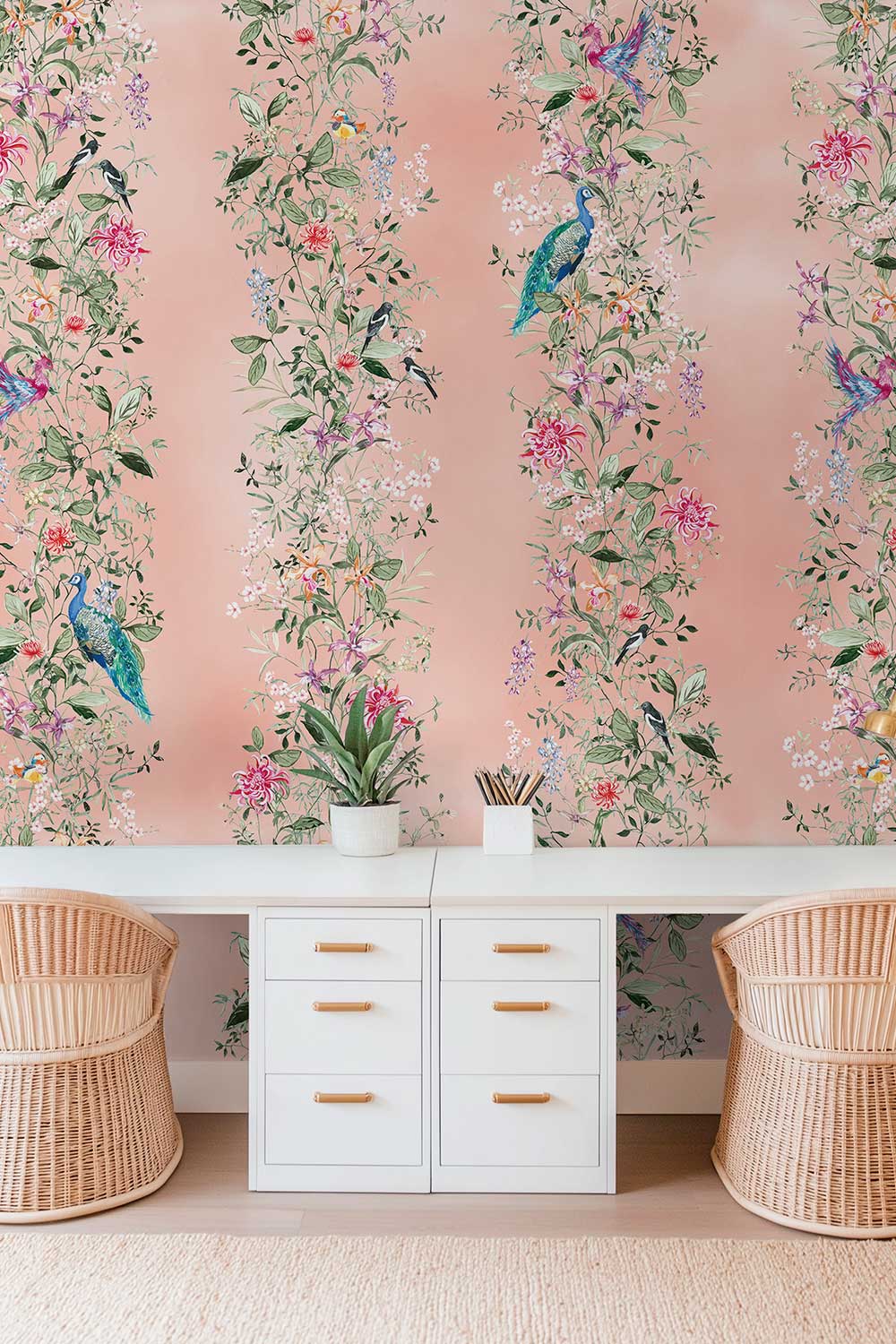 A styled interior scene featuring vertical floral wallpaper with peacocks and colorful blossoms on a soft blush pink background, behind a white desk with drawers and two woven rattan chairs. Hand-painted Mahjong Garden Stripe wallpaper featuring watercolor chinoiserie florals, peacocks, birds, bamboo, and botanical stripes on an blush pink coral background