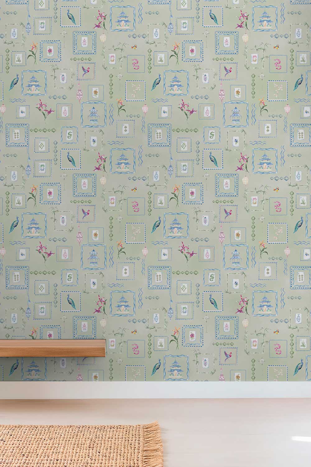 A close-up of a wallpaper pattern featuring a light sage green background with a repeating motif of blue-framed cartouches, birds, and small floral details, shown on a wall above a white baseboard and a woven rug. Hand-painted watercolor Mahjong Tiles wallpaper featuring mahjong tiles, pagodas, bamboo, birds, ginger jars, lanterns, and chinoiserie artwork on a soft green background printed on an all-natural and eco-friendly grasscloth wall-covering.