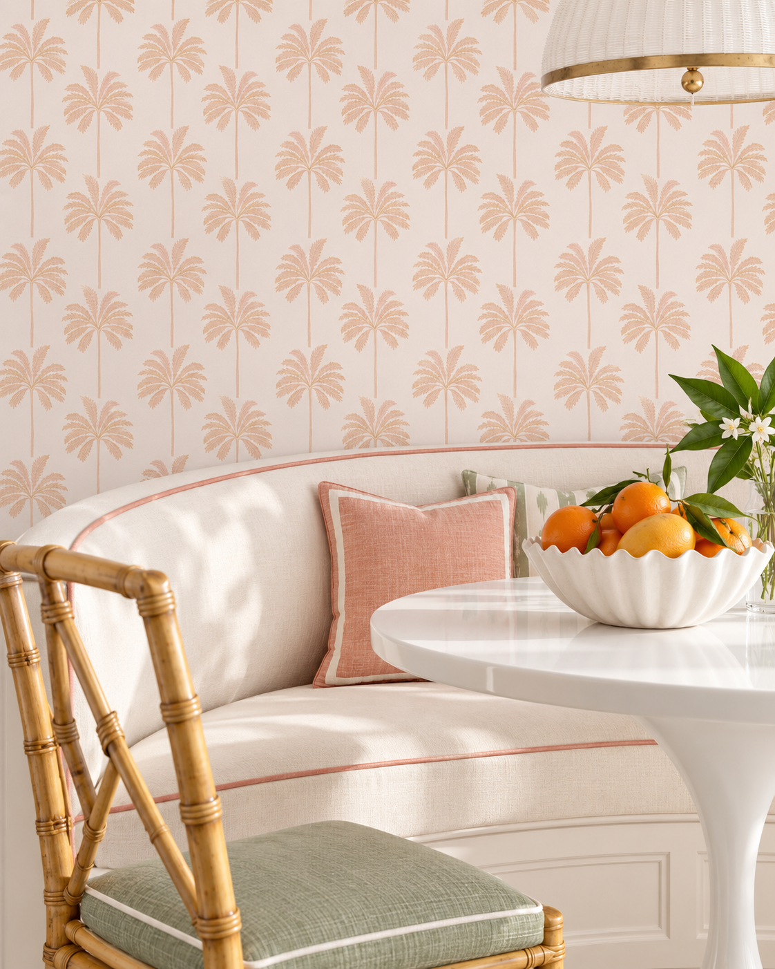 A styled interior corner featuring a curved cream upholstered bench with a pink pillow, a white round table with a bowl of oranges, a bamboo chair with a green cushion, and wallpaper with a repeating beige palm leaf pattern on a light background.