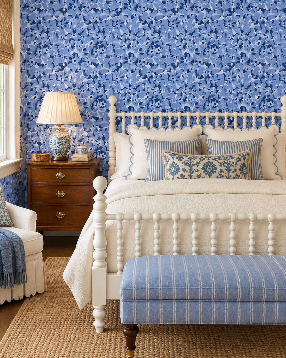 A beautifully styled bedroom featuring a white wooden spindle bed dressed in cream bedding, a blue and white patterned bench at the foot, a wooden nightstand with a lamp, and a blue floral wallpapered wall. cozy cottage guest bedroom with vintage inspired blue floral design printed on luxurious all-natural grass cloth wall-covering