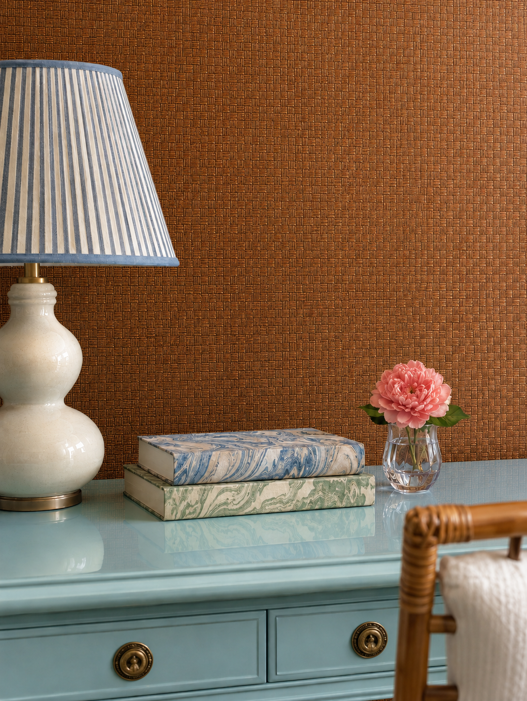 A styled interior scene showing a light blue dresser against a textured rust-brown wall, with a white table lamp, stacked books, and a small vase of pink flowers. All-Natural Square Weave Paperweave Wallpaper – Eco-Friendly Dark Walnut brown color, eco-friendly wall-covering