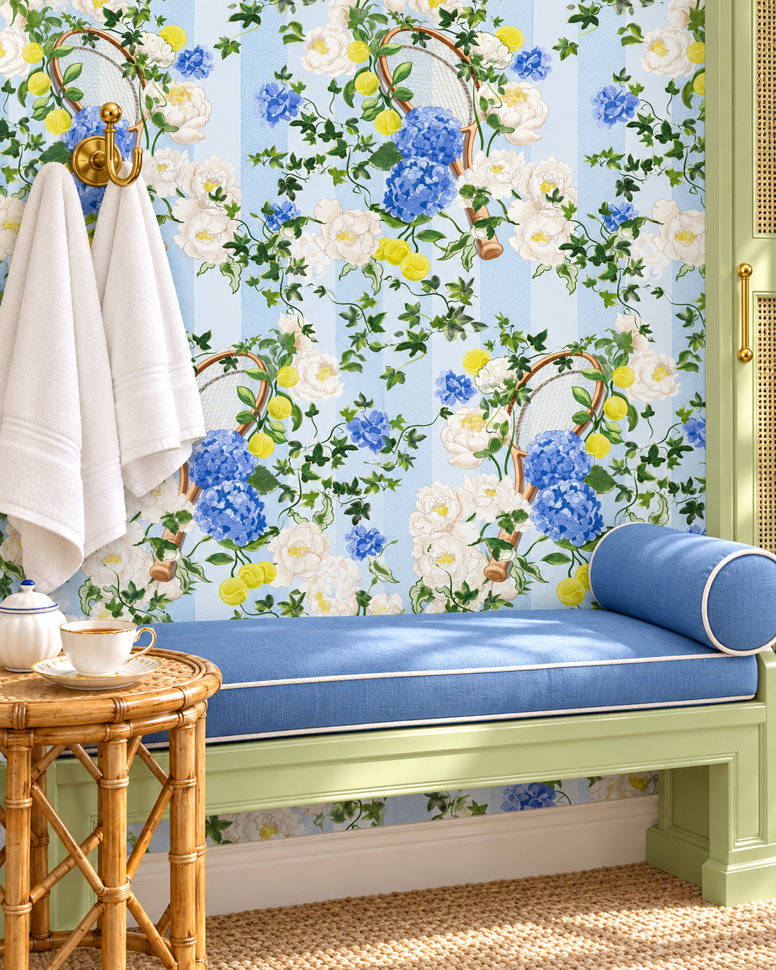 A styled interior scene featuring a light blue bench with a blue cushion, white towels hanging on a gold hook, and a wall covered in floral wallpaper with blue, white, and yellow flowers on a pale blue background. Hand-painted botanical stripe wallpaper with hydrangeas, peonies, tennis rackets, and tennis balls on a classic matte finish