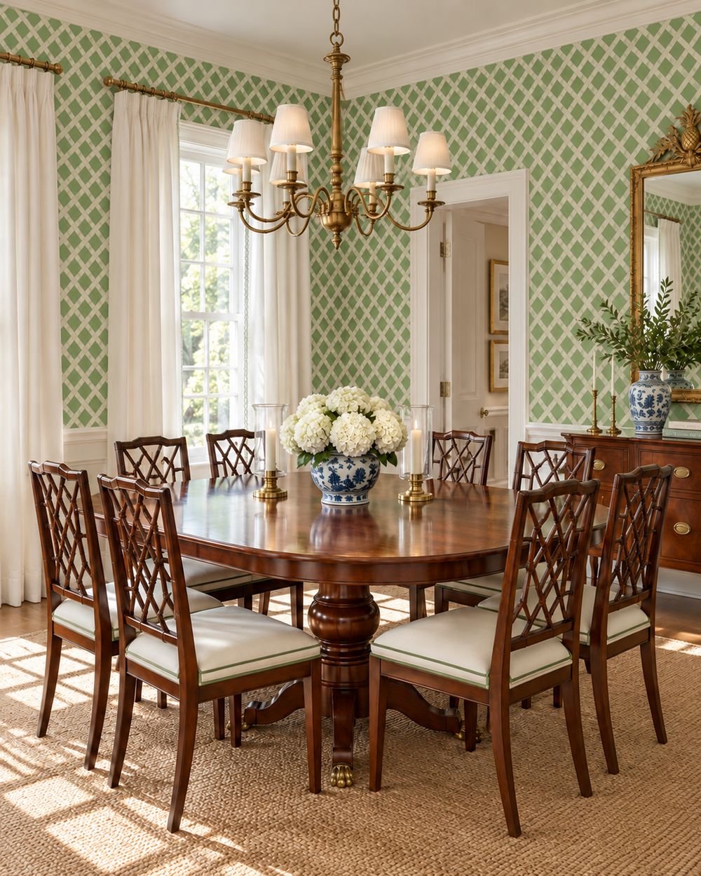 A classic dining room featuring a round dark wood dining table surrounded by eight matching wooden chairs with cream upholstered seats. The room is decorated with green and white patterned wallpaper, white curtains, a brass chandelier, and a large mirror on a wooden console table.