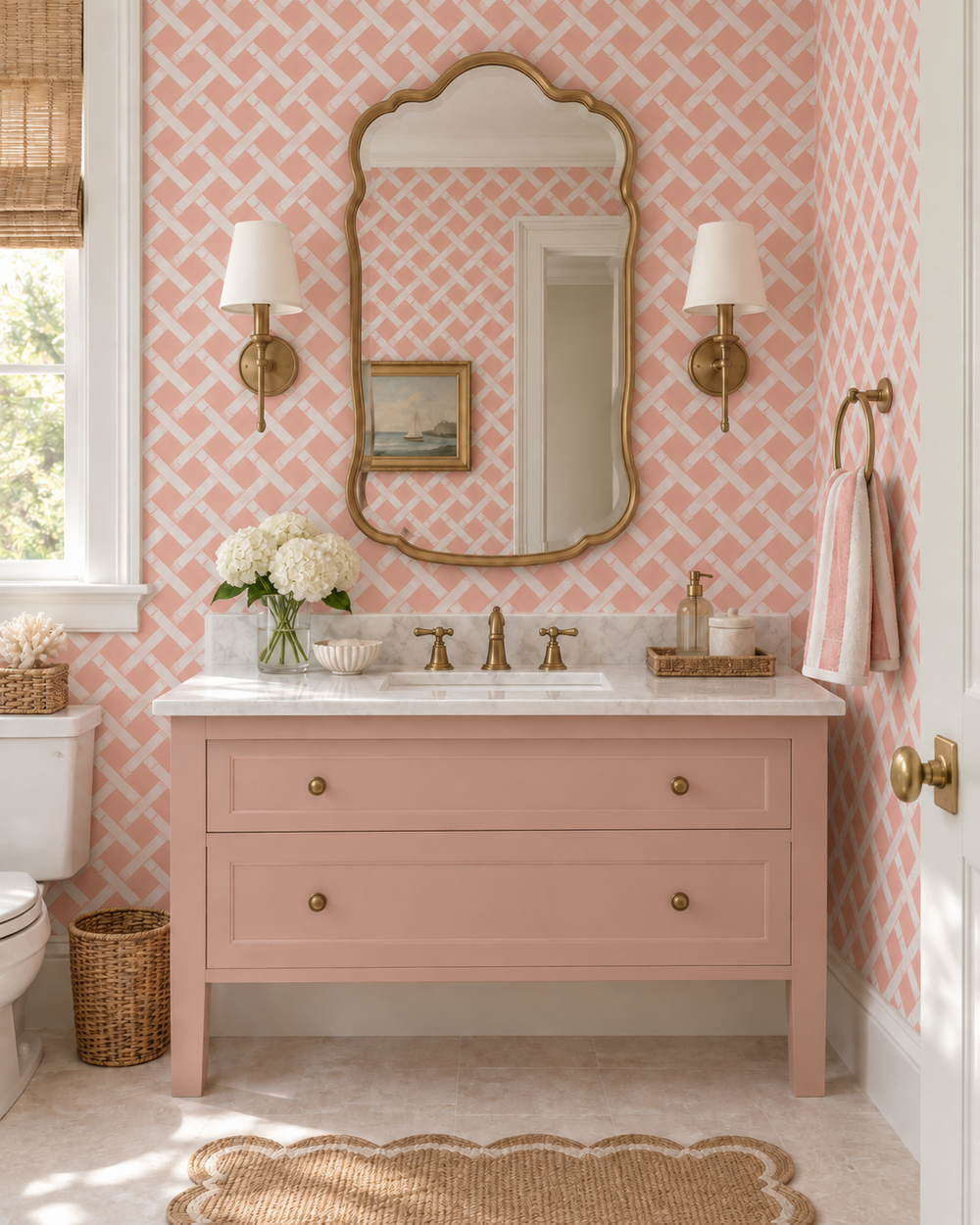 A pink wooden bathroom vanity with a white marble countertop, gold faucet and knobs, set against pink patterned wallpaper. An ornate gold-framed mirror hangs above, flanked by two gold wall sconces.