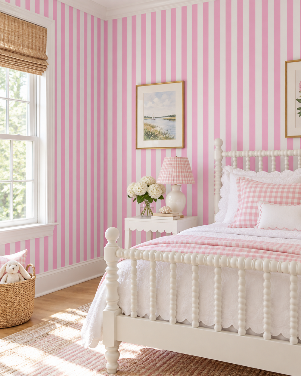 A styled bedroom featuring pink and white vertical striped wallpaper, a white wooden bed with pink gingham bedding, a white nightstand with a lamp and flowers, and a window with a woven shade.