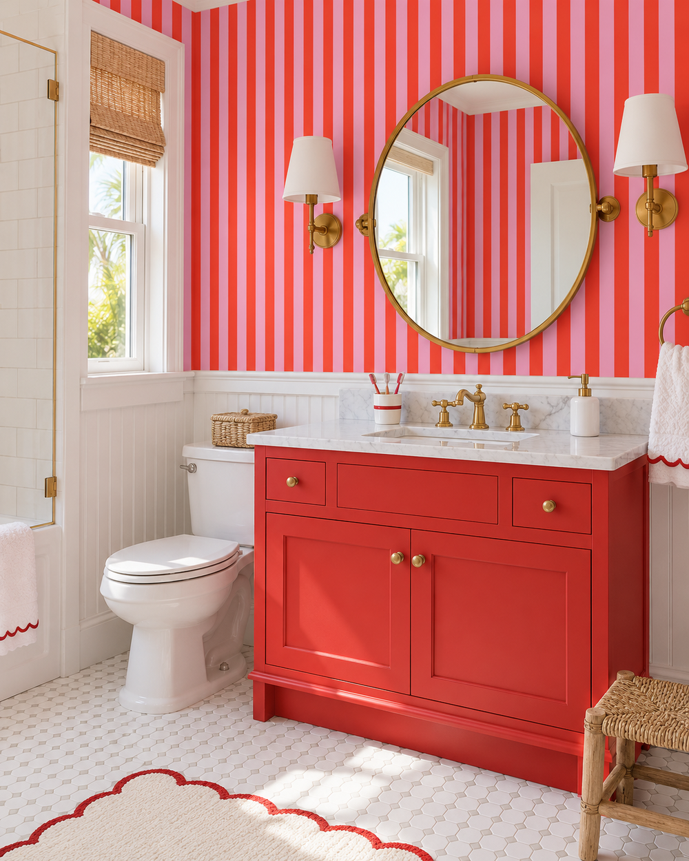 A bright bathroom featuring bold coral pink and white vertical striped wallpaper, a bright red vanity cabinet with brass hardware, a white marble countertop, a large round gold-framed mirror, and a white toilet.