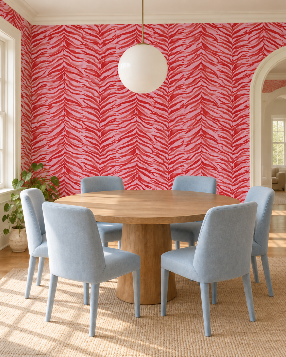 A styled dining room featuring a round wooden table surrounded by six light blue upholstered chairs, set against a bold red and white zebra-print wallpaper wall. A white globe pendant light hangs from the ceiling, and a potted plant sits on the floor near the window. feminine pink and red watercolor zebra stripe printed on commercial grade water and stain resistant performance vinyl wall-covering, great for bathrooms, kitchens and commercial spaces