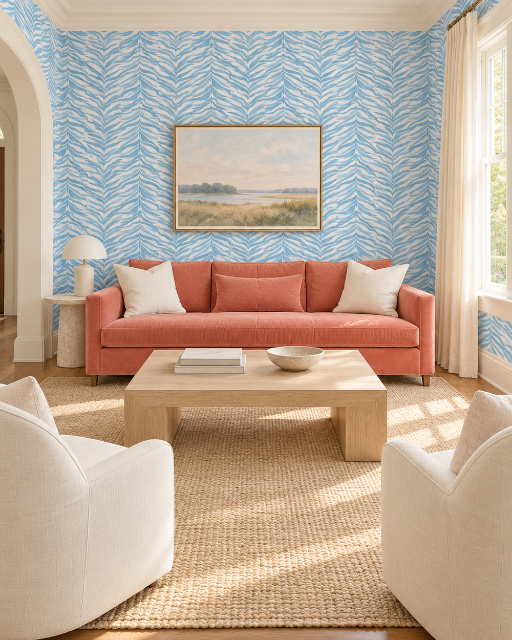 A styled living room featuring a coral pink sofa with white pillows, set against a wall with light blue and white leaf-patterned wallpaper. A light wood coffee table sits on a beige woven rug, with two cream armchairs in the foreground and a framed landscape painting on the wall.