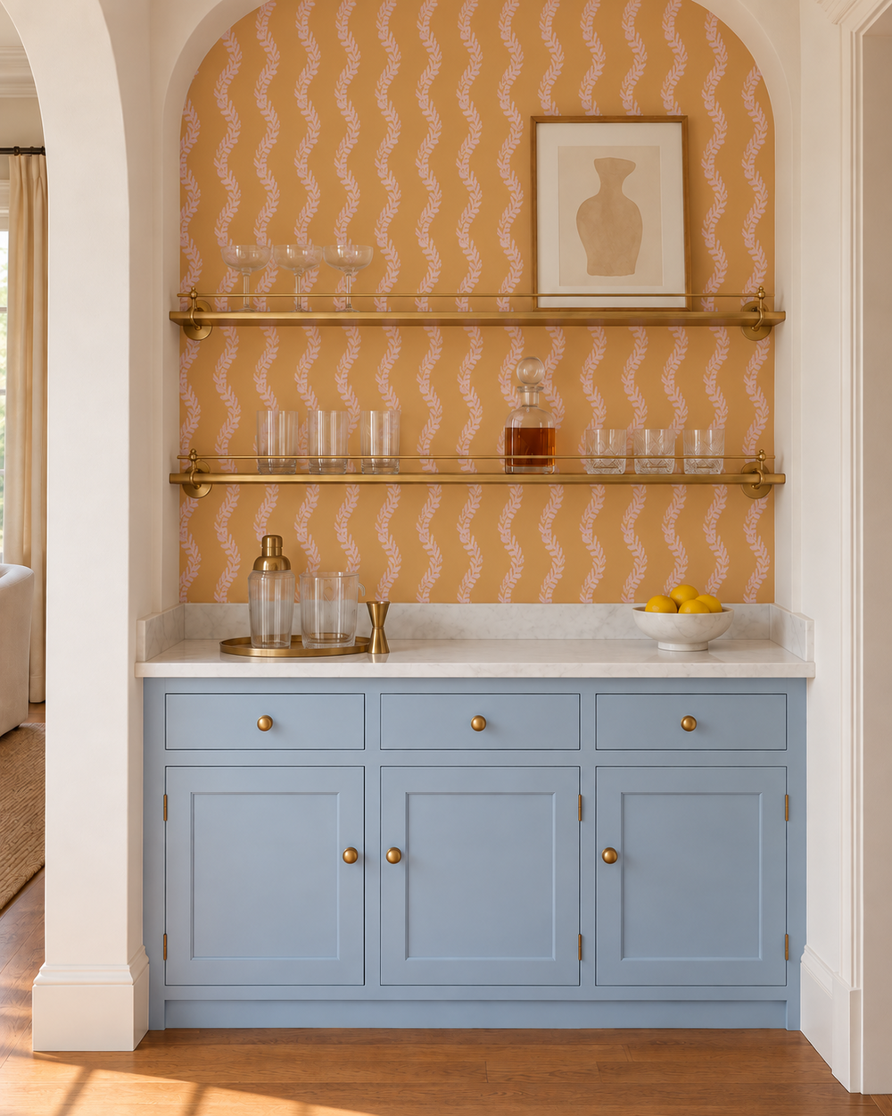 A built-in bar cabinet in a home interior, featuring light blue shaker-style doors with brass knobs, a white marble countertop, two brass floating shelves, and a peach patterned wallpaper backdrop.