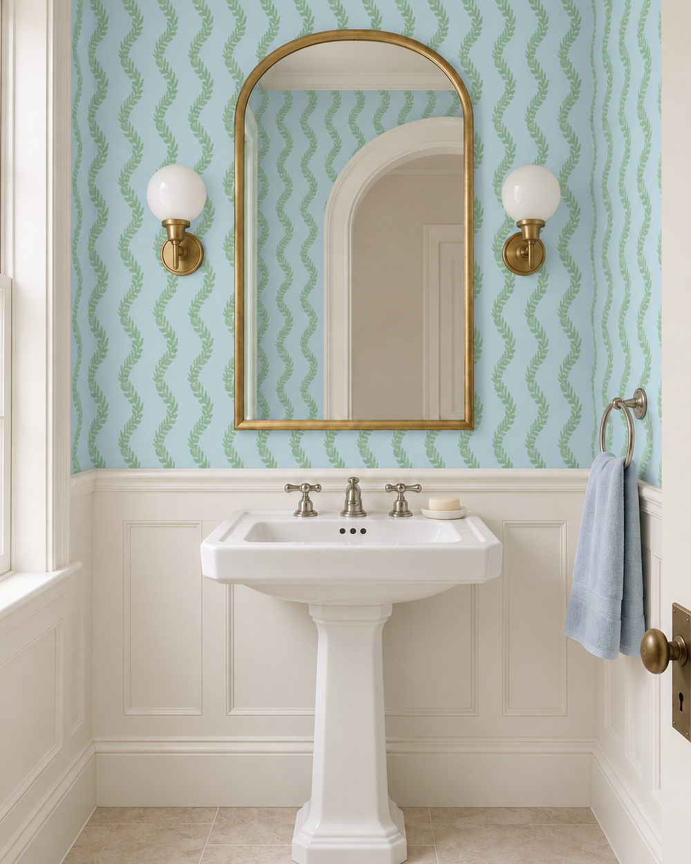 A styled bathroom interior featuring a white pedestal sink, a gold-framed arched mirror, and light blue wallpaper with a green wavy pattern. The wall is also decorated with white wainscoting, two gold wall sconces, a blue towel on a ring, and a window on the left.